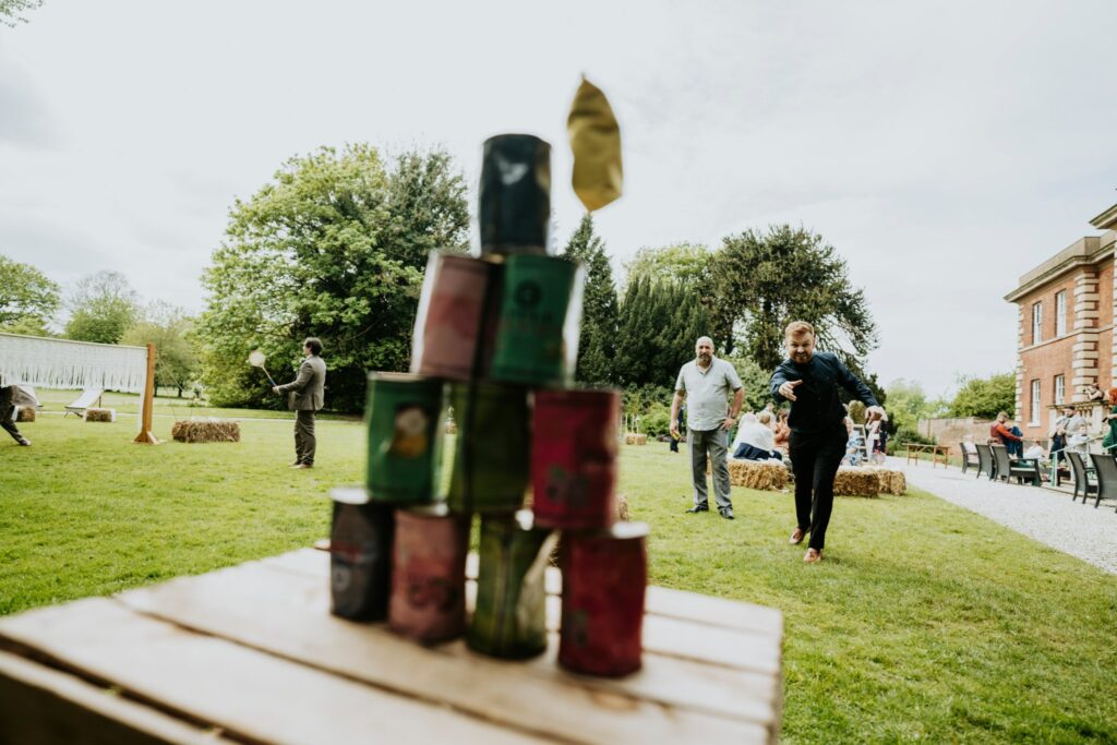 Wedding guests playing a game of Tin Can Alley on a wedding day at Hirst Priory in Crowle, Lincolnshire with other guests playing a game of Badminton in the background with a luxury Macrame Badminton Net | Luxury Garden Lawns Games Hire in Lincolnshire