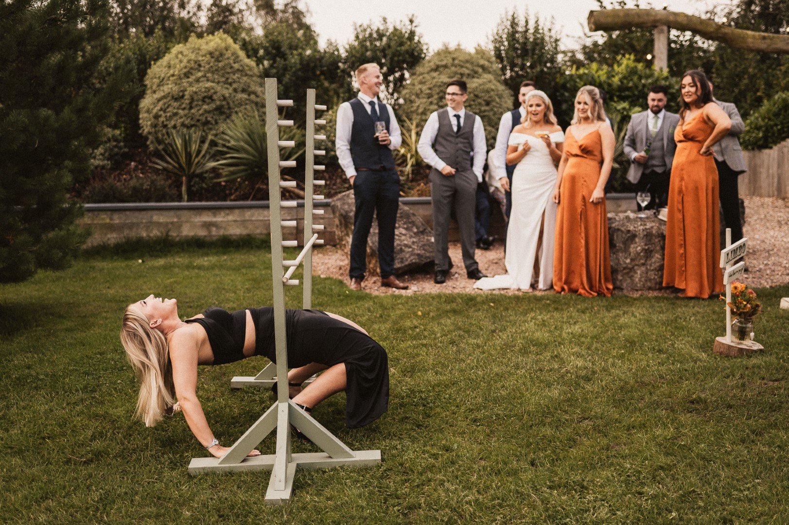 Wedding guests are stood watching as a woman plays Limbo and is falling backwards.
